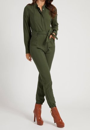 Olijfgroene jumpsuit met lange mouwen, knoopsluiting aan de voorkant, taillekoord en taps toelopende pijpen, gecombineerd met bruine hoge hakken met krokodillenprint.