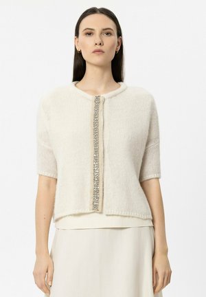 Frau trägt eine cremefarbene, kurzärmelige Strickjacke mit einer vertikalen Linie dekorativer silberner Verzierungen auf der Vorderseite.