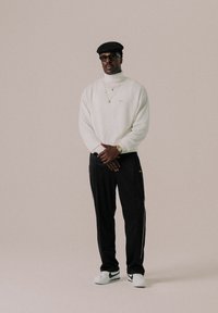 Maglione bianco a collo alto, pantaloni da jogging neri con strisce laterali, berretto nero, occhiali trasparenti e sneakers bianche con logo nero.