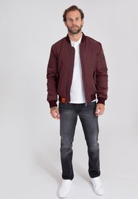 Giacca bomber borgogna con polsini a costine, zip frontale e tasche laterali; indossata sopra una polo bianca e abbinata a jeans grigi e sneakers bianche.