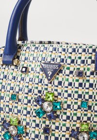 Woven handbag with blue handles, featuring a textured fabric in navy and cream, adorned with colorful jeweled accents and a logo plaque.
