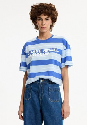 Mujer con cabello rizado lleva una camiseta de rayas azul y azul claro con texto "CHASE SMALL WONDERS" y vaqueros azules de talle alto con bolsillos delanteros.
