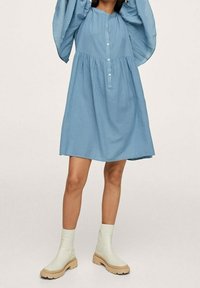 Robe à carreaux bleus avec des manches bouffantes, col boutonné à l'avant et taille froncée. Associée à des bottines crème à la cheville avec une semelle texturée.