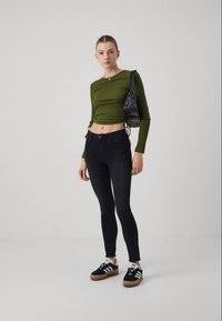 Camiseta verde de manga larga, recortada con fruncidos laterales, combinada con vaqueros negros ajustados y zapatillas negras con rayas blancas y suela de goma.