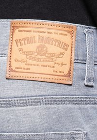 Etiqueta de cuero en denim azul claro con "PETROL INDUSTRIES" grabado. Presenta detalles de costura y una superficie texturizada.