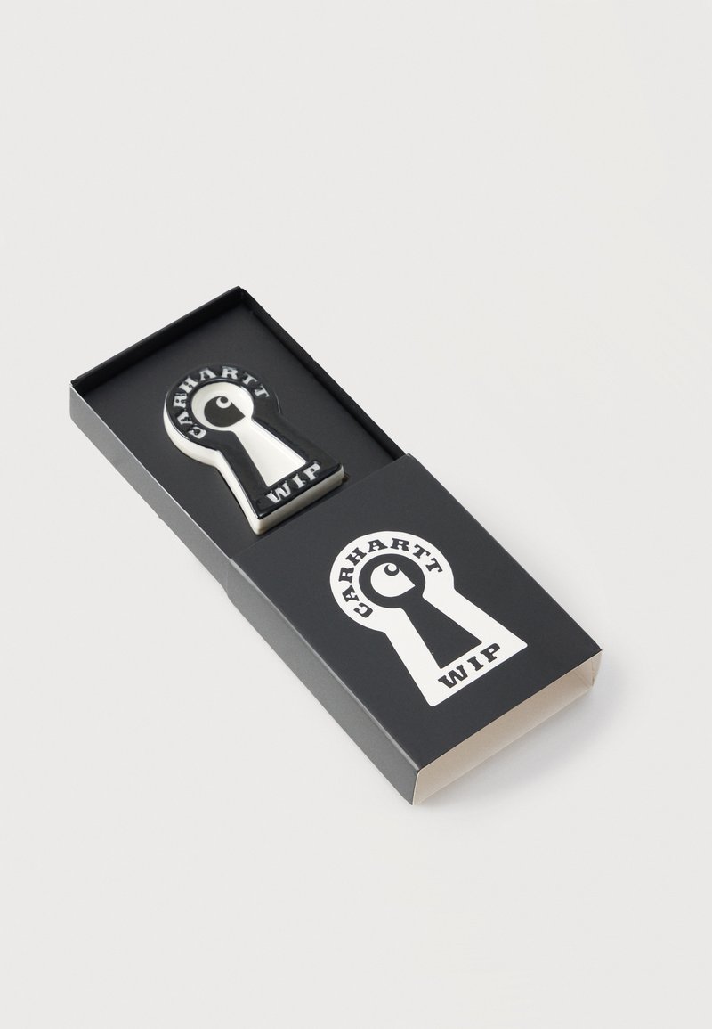 Schwarz-weiße Carhartt WIP Anstecknadel in Schlüssellochform in passender schwarzer Box mit gleichem Schlüsselloch-Logo auf dem Deckel.