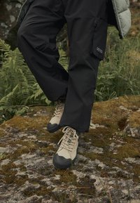 Beige Wanderschuhe mit schwarzen Gummispitzen, strukturierten Schnürsenkeln und dunkler Hose mit einem Reißverschluss-Cargotasche, auf einem steinigen, moosbedeckten Boden.