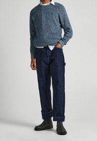 Muž v modrém pleteném svetru přes bílou košili, tmavě modrých vyhrnutých kalhotách a černých Chelsea botách, stojící s jednou rukou v kapse.