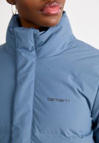 Lyseblå hættetrøje med lynlås og høj krave, ensartet design og Carhartt-logo i mørk tryk på brystet. Glat tekstur.