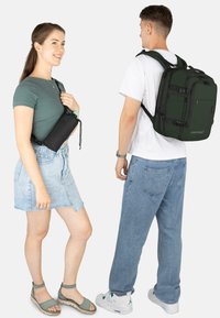 Schwarze Umhängetasche und grüner Rucksack mit mehreren Fächern. Beide bestehen aus strapazierfähigem Stoff; die Tasche verfügt über einen Kordelzugverschluss.