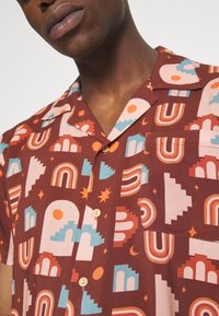 Lyhythihainen, ruskea kuvioitu paita, jossa on geometrisia muotoja, holveja ja tähtiä vaaleanpunaisessa, oranssissa ja teal-värissä. Sisältää rintataskun.