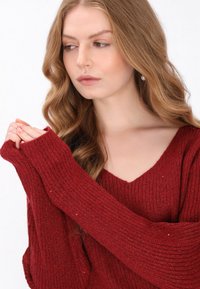 Piros bordás pulóver mély V-nyakkivágással és hosszú ujjakkal; texturált anyag jellemzi, diszkrét csillámszínekkel átszövegezve.