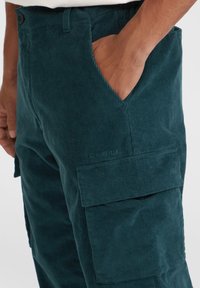 Teal corduroy cargobroek met een zijzak, textuurstof en geborduurd logo-detail op de bovenbenen.