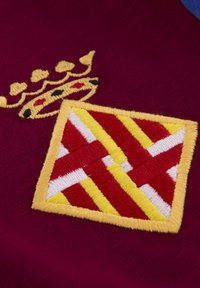 Geborduurd patch op bordeaux kleurige stof met een gouden kroon en een diamanten ontwerp met rode, gele en witte patronen.