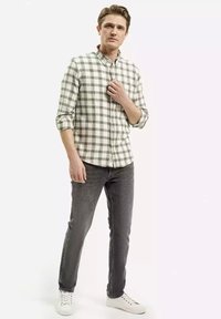 Męska koszula zapinana na guziki w biało-zieloną kratę z podwiniętymi rękawami, zestawiona z ciemnoszarymi dżinsami i białymi trampkami.