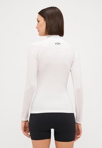 Bijela dugouha rash guard majica s visokim izrezom i ravnim šavovima, u kombinaciji s crnim kratkim hlačama. Na leđima se nalazi logotip brenda.