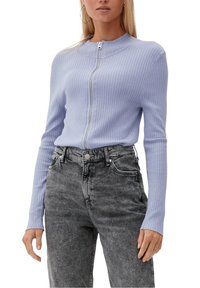 Jasnoniebieski sweter z prążkowanego materiału z suwakiem z przodu i długimi rękawami, noszony z szarymi jeansami z wysokim stanem. Tkanina wydaje się miękka i teksturowana.