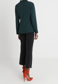 Tmavě zelená halenka s dlouhým rukávem a vysokým límcem, kombinovaná s černými zkrácenými džínami a oranžovými lodičkami s pásky kolem kotníků.