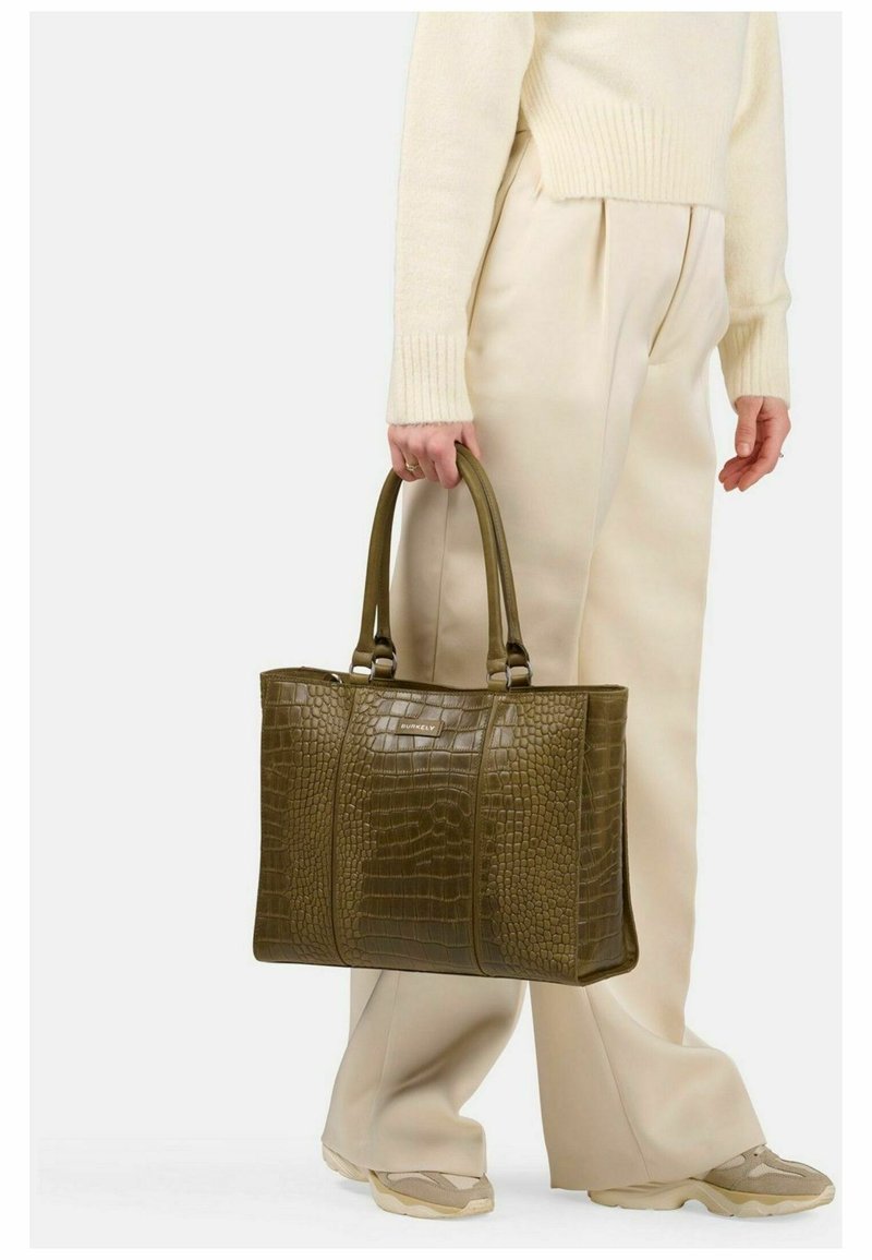Borsa tote in pelle stampata a coccodrillo verde oliva con doppi manici e una piccola toppa con logo, abbinata a pantaloni plissettati color crema e scarpe chiare.