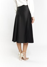 Schwarzer Satin-Midirock mit glatter Textur, ausgestelltem Design und einem dezenten seitlichen Schlitz. Kombiniert mit goldenen High-Heels.