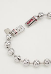 Bracciale in metallo tono argento con perle sferiche e chiusura con il marchio "HUGO" in rosso. Texture liscia e design moderno.