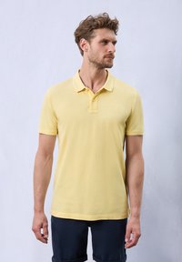 Gul bomullspolo med krage och tre knappar, med korta ärmar och en diskret logodetalj på bröstet.