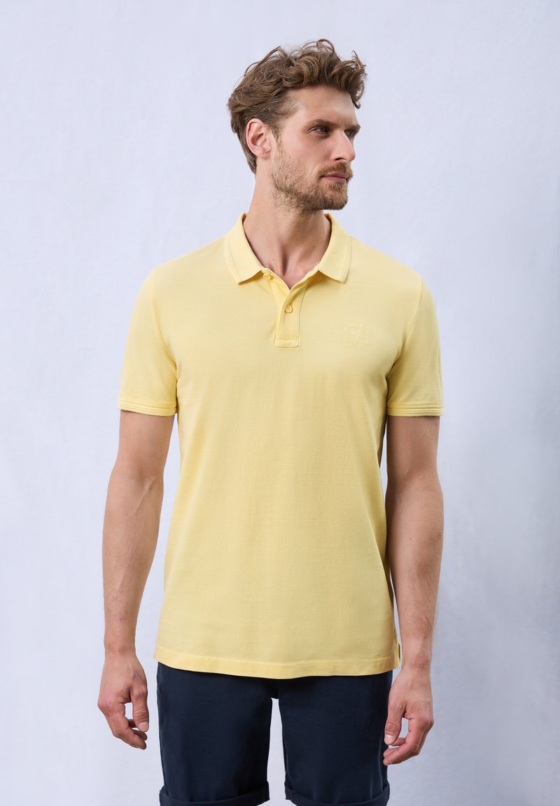 Gul bomullspolo med krage och tre knappar, med korta ärmar och en diskret logodetalj på bröstet.