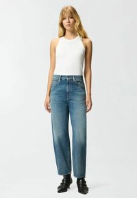 Top blanco sin mangas combinado con vaqueros de pierna ancha de cintura alta en azul claro. Los vaqueros tienen un acabado desgastado, y el modelo lleva botas negras hasta el tobillo con detalles metálicos.