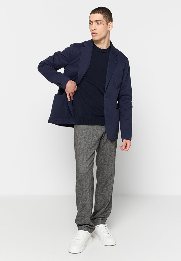 Blazer navy sur un pull sombre, associé à un pantalon gris texturé et des baskets blanches. Design élégant avec des poches sur le blazer.