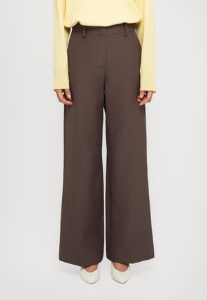 Pantaloni larghi marroni con passanti per cintura, frontale liscio e tessuto leggermente strutturato, abbinati a un top giallo chiaro a maniche lunghe e tacchi bianchi.