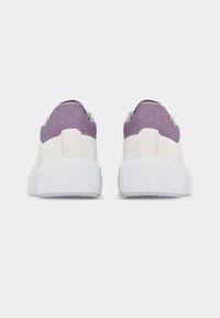 Ζευγάρι λευκών αθλητικών παπουτσιών με χοντρές λευκές σόλες και μοβ σουέντ περιλαίμια στη φτέρνα, εμφανιζόμενα από πίσω σε απλό ανοιχτόχρωμο φόντο.