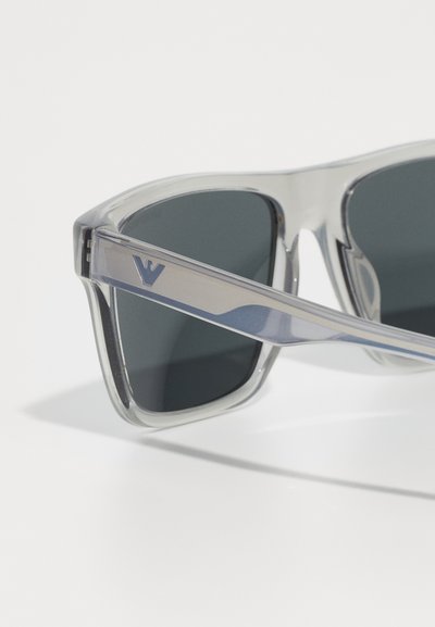 Ochelari de soare cu o ramă gri transparent, lentile închise la culoare și un accent argintiu pe braț cu un logo. Textură netedă, lucioasă.