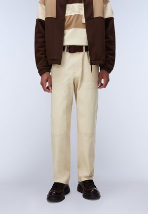 Mann trägt cremefarbene Hosen mit aufgesetzten Fronttaschen, dunkelbraunen Gürtel, dunkelbraune Schuhe und eine braun-beige Jacke mit Reißverschluss.