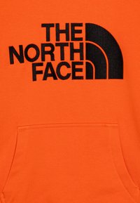 Felpa arancione con un grande logo nero di "THE NORTH FACE" e una tasca frontale. Texture liscia con un design classico a collo rotondo.
