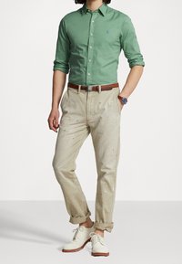 Zielona koszula zapinana na guziki, beżowe spodnie z plamami farby, brązowy skórzany pasek i jasne buty z sznurówkami. Prezentowany strój casualowy.