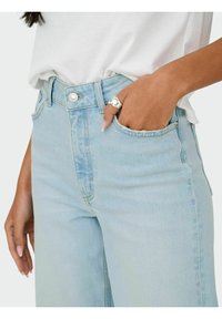 Ανοιχτού μπλε ψηλόμεσες τζιν με ελαφριά εφέ ξεθωριάσματος. Διαθέτουν φερμουάρ, κουμπί κλεισίματος και τσέπες, συνδυασμένες με λευκό μπλουζάκι.