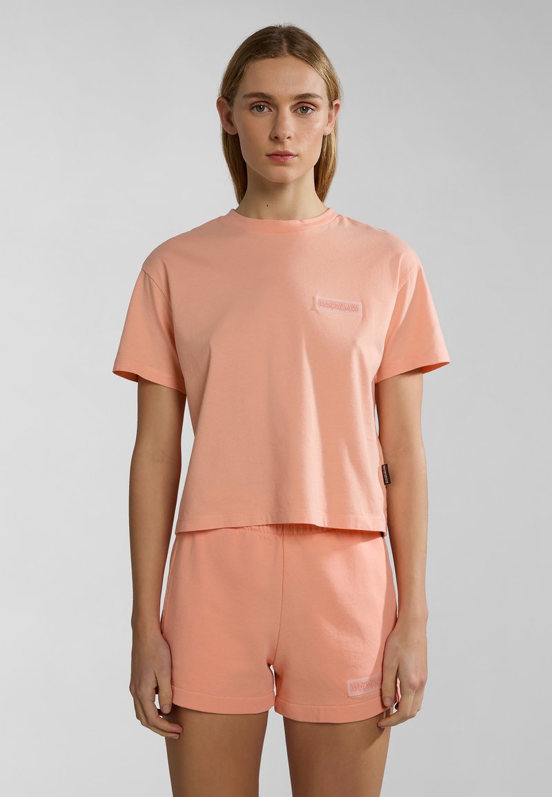 Camiseta de algodón de color melocotón con mangas cortas y un corte cuadrado, acompañada de pantalones cortos a juego. Presenta un pequeño logo bordado en ambos artículos.