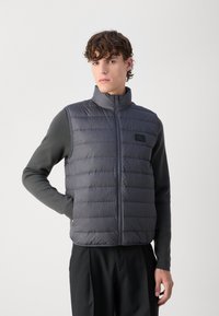 Gepolsterte graue Weste mit hohem Kragen, Reißverschluss und kleinem Logo, getragen über einem enganliegenden Langarmshirt mit dunklen Ärmeln und schwarzen Hosen.