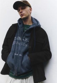 Černá vnější bunda, modrá mikina s kapucí s nápisem, zelené pruhované tričko pod ní, šedé tepláky a hnědá čepice. Texturované materiály, vrstvené oblečení.