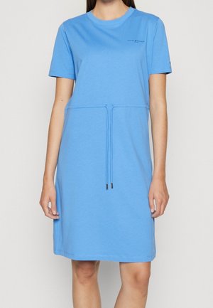 Woman wearing a light blue short-sleeve dress with a drawstring waist and small logo on the chest against a plain background.