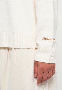 Suéter de punto crema con puños y dobladillo acanalados. Presenta texto bordado en marrón claro en la manga. Textura suave, ajuste relajado.