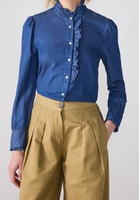 Jeansskjorta med rynkad front och hög krage, knäppt, i kombination med högmidjade khakibyxor med veck och knappstängning.