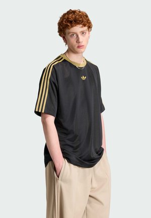 Mladá osoba s kudrnatými rezavými vlasy, oblečená v černém tričku Adidas se zlatými pruhy a béžovými kalhotami, ruce v kapsách, neutrální výraz.
