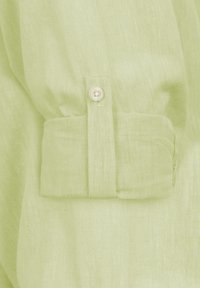 Camisa de lino verde claro con puños abotonados que tienen un único botón blanco y una tela suave y texturizada. Los detalles incluyen un diseño de puño doblado.