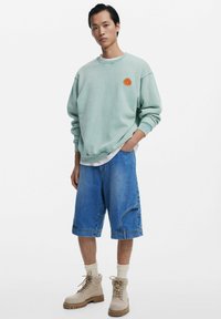 Sweatshirt vert menthe avec un graphisme orange, associé à un short en denim bleu et des bottes à lacets beiges. La tenue présente une coupe décontractée et confortable.