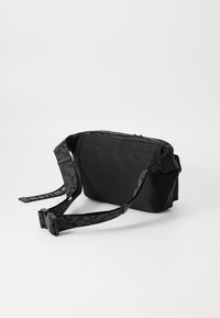 Borsa nera in nylon di forma rettangolare, con tracolla regolabile a fantasia, chiusura con zip e tasche laterali piatte. Il testo sulla tracolla recita "marimekko."