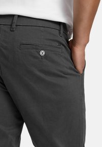 Person wearing dark gray pants with a back buttoned pocket and a hand in the front pocket, paired with a white shirt.