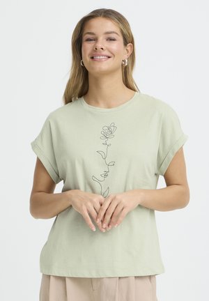 Femme souriante aux longs cheveux portant des boucles d'oreilles en anneau, un t-shirt vert pâle à manches retroussées avec un design continu de ligne de rose noire, pantalon beige.