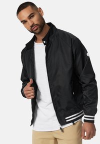 Chaqueta bomber ligera negra con cuello, cierre de cremallera y puños a rayas. Cuenta con bolsillos laterales y un detalle de logo en la manga.