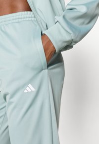 Ανοιχτό μπλε παντελόνια φόρμας με υφή, ελαστική μέση, πλαϊνή τσέπη και λευκό λογότυπο Adidas στο κάτω αριστερό πόδι.